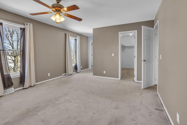 unfurnished bedroom with multiple windows, a walk in closet, light colored carpet, and ceiling fan