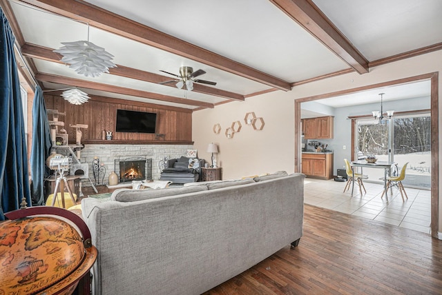 living room with beamed ceiling, a stone fireplace, ceiling fan with notable chandelier, and light wood-type flooring