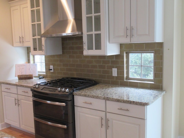 kitchen with wall chimney exhaust hood, light stone counters, tasteful backsplash, range with two ovens, and white cabinets