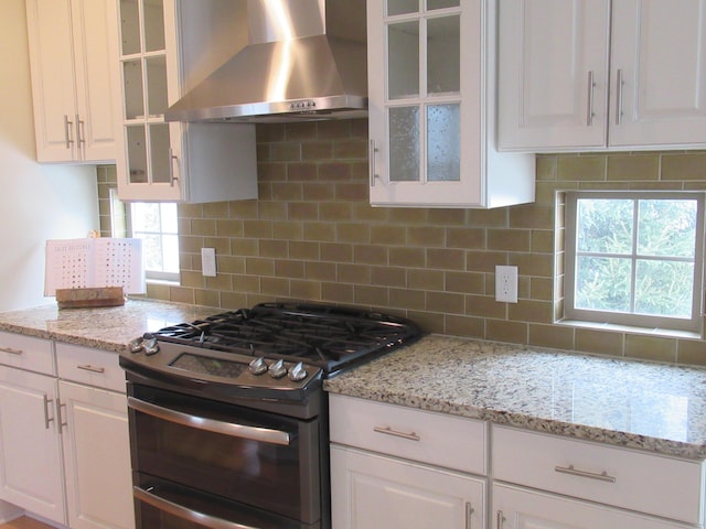 kitchen featuring backsplash, light stone countertops, white cabinets, wall chimney exhaust hood, and range with two ovens