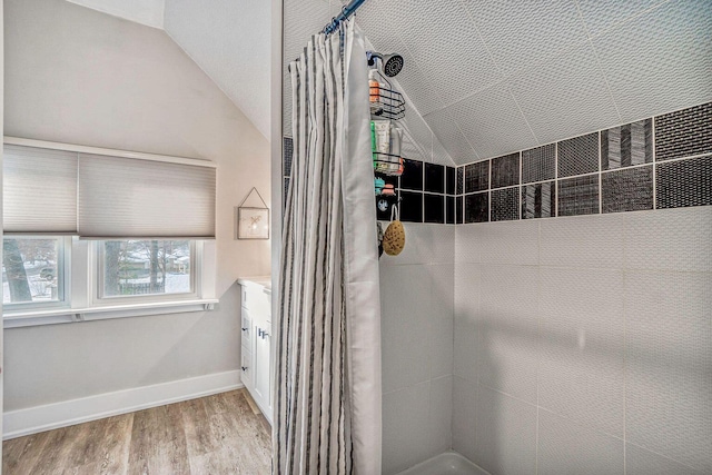 full bath featuring wood finished floors, vanity, baseboards, vaulted ceiling, and a tile shower