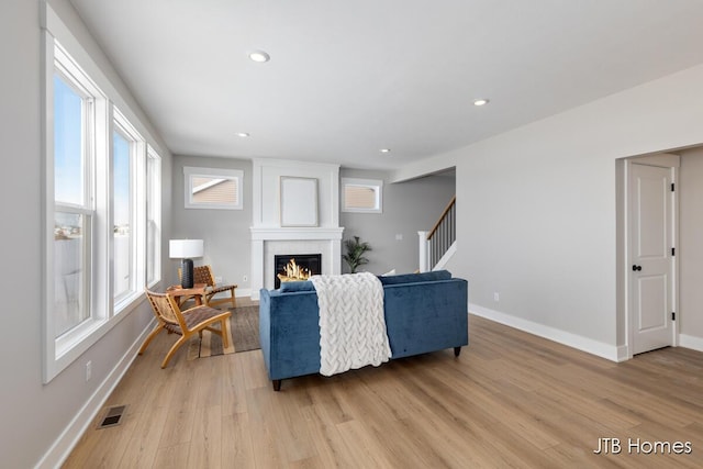 living room featuring a warm lit fireplace, light wood finished floors, baseboards, visible vents, and stairs