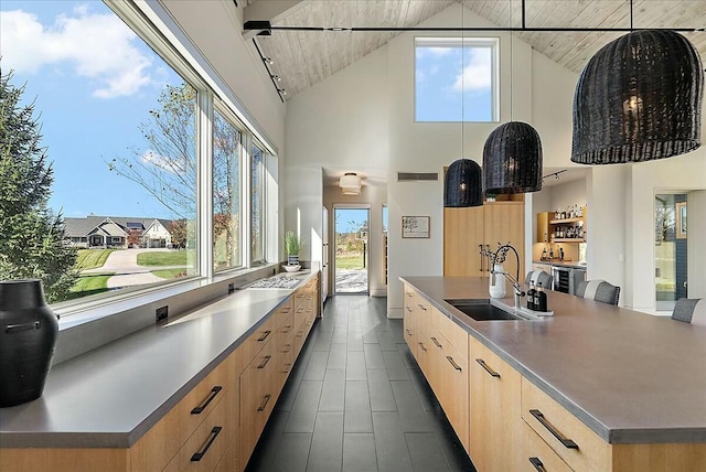 kitchen with light brown cabinetry, sink, high vaulted ceiling, hanging light fixtures, and a kitchen island with sink