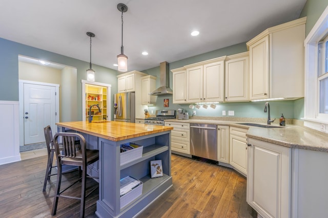 kitchen with a center island with sink, stainless steel appliances, decorative light fixtures, wall chimney range hood, and wooden counters