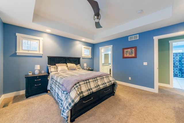 bedroom featuring a raised ceiling, ensuite bathroom, light carpet, and ceiling fan