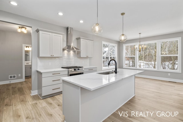 kitchen featuring a sink, wall chimney range hood, stainless steel range with gas cooktop, a center island with sink, and light stone countertops