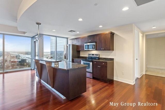 kitchen featuring dark wood-type flooring, appliances with stainless steel finishes, hanging light fixtures, dark brown cabinets, and a center island with sink