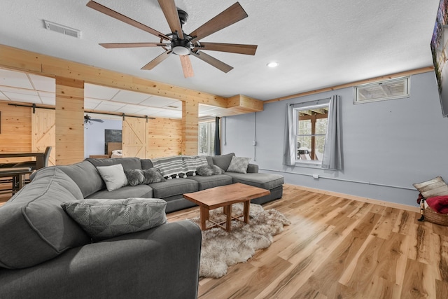 living room featuring wooden walls, ceiling fan, light hardwood / wood-style floors, a barn door, and a textured ceiling