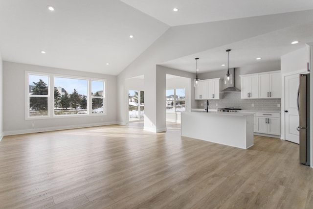 kitchen with wall chimney range hood, stainless steel refrigerator, an island with sink, white cabinets, and decorative light fixtures