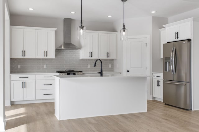 kitchen featuring wall chimney exhaust hood, white cabinetry, decorative light fixtures, stainless steel fridge with ice dispenser, and a kitchen island with sink