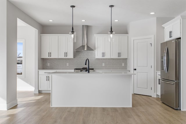 kitchen with decorative light fixtures, white cabinetry, stainless steel fridge with ice dispenser, a center island with sink, and wall chimney exhaust hood