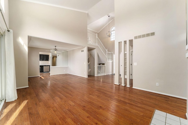 unfurnished living room featuring wood-type flooring, a towering ceiling, ceiling fan with notable chandelier, and ornamental molding