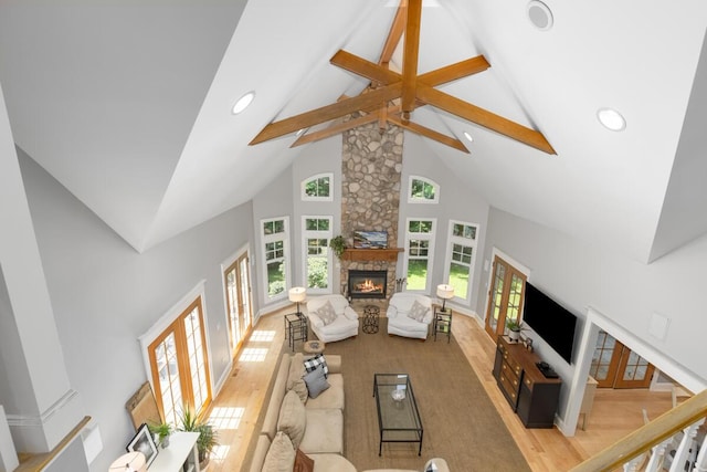 living room featuring french doors, a stone fireplace, high vaulted ceiling, light hardwood / wood-style flooring, and beam ceiling