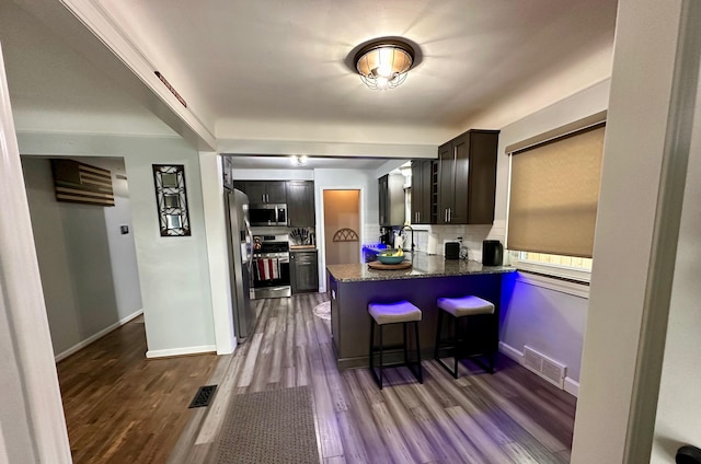 kitchen featuring dark wood-type flooring, appliances with stainless steel finishes, dark brown cabinets, tasteful backsplash, and a kitchen bar