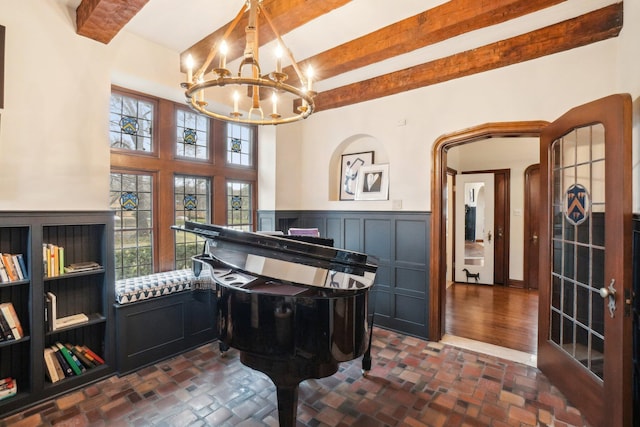living area featuring beamed ceiling, brick floor, a chandelier, and wainscoting