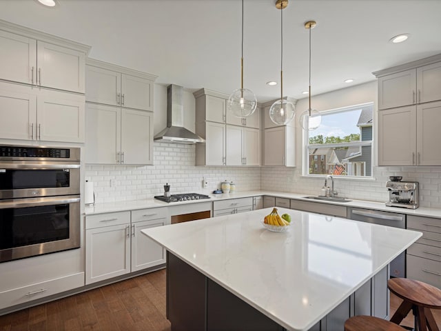 kitchen featuring a kitchen island, a sink, hanging light fixtures, wall chimney range hood, and appliances with stainless steel finishes