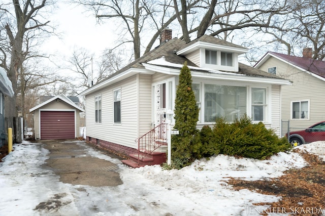 snow covered property with a detached garage, a chimney, a shingled roof, concrete driveway, and an outdoor structure