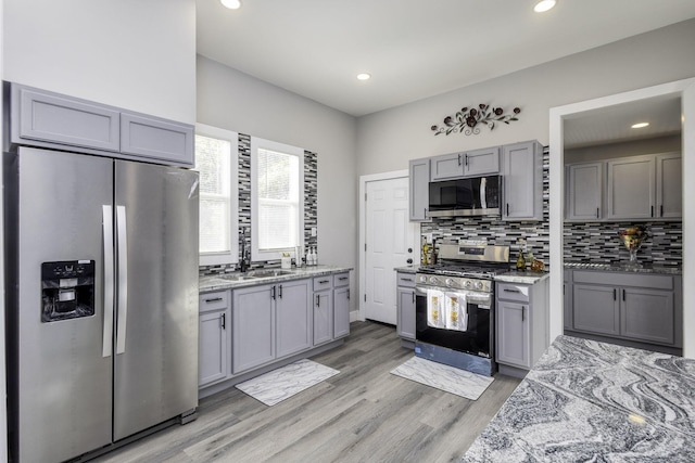 kitchen with light stone counters, gray cabinets, light wood-style flooring, backsplash, and appliances with stainless steel finishes