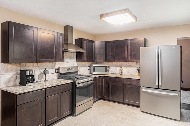 kitchen featuring light stone counters, decorative backsplash, appliances with stainless steel finishes, dark brown cabinets, and wall chimney exhaust hood