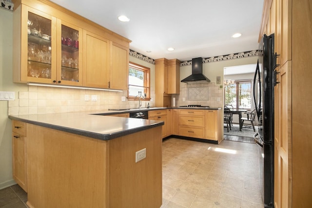 kitchen featuring dark countertops, light brown cabinetry, glass insert cabinets, a sink, and wall chimney exhaust hood