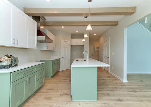 kitchen featuring decorative light fixtures, light countertops, a kitchen island with sink, beamed ceiling, and wall chimney exhaust hood