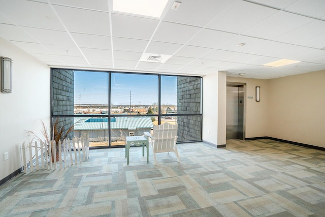 empty room with a paneled ceiling, elevator, carpet flooring, a wall of windows, and baseboards