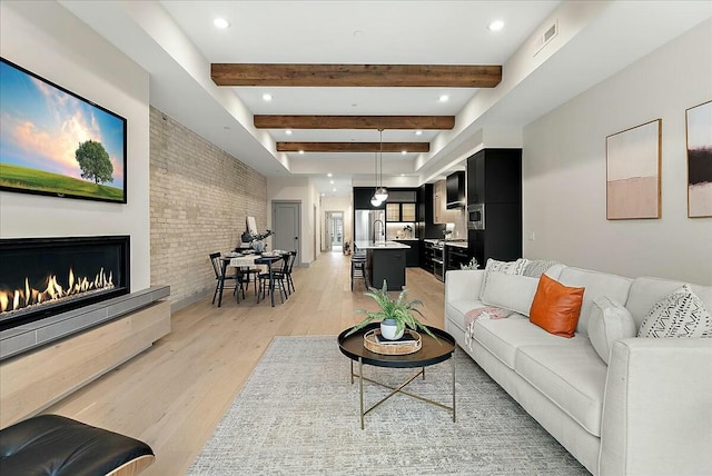 living room with a warm lit fireplace, brick wall, beamed ceiling, light wood-style floors, and recessed lighting