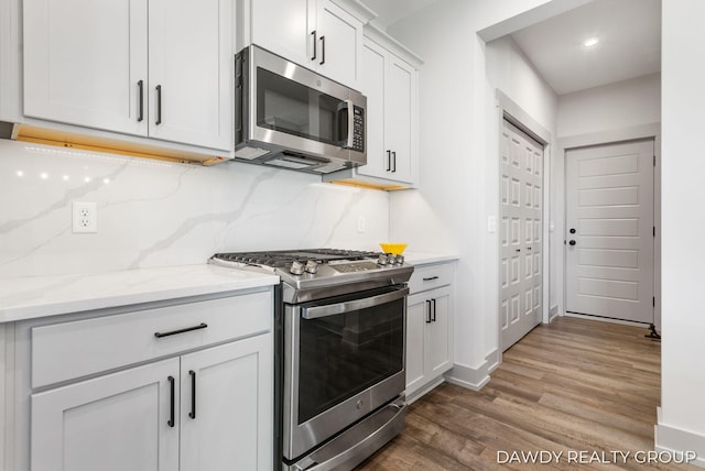 kitchen with white cabinets, light wood-style flooring, appliances with stainless steel finishes, light stone counters, and backsplash