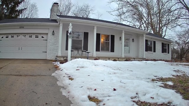 single story home with driveway, a chimney, an attached garage, covered porch, and brick siding