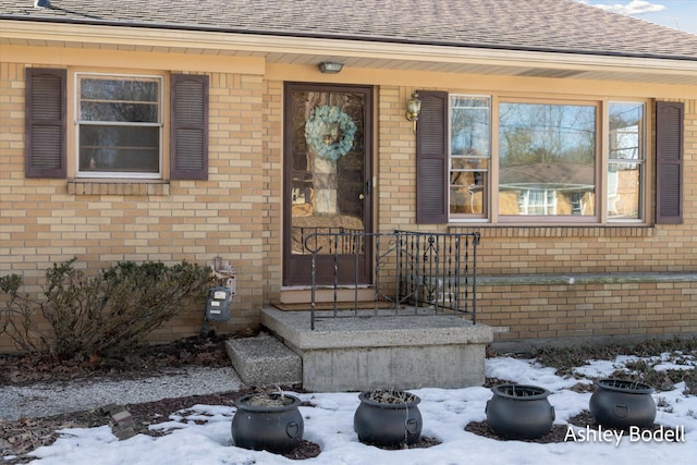 snow covered property entrance with a shingled roof and brick siding