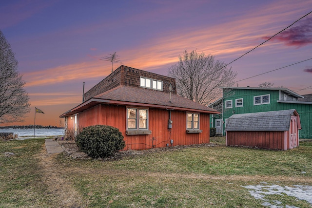 back of property featuring an outbuilding, a shingled roof, a water view, a lawn, and a shed