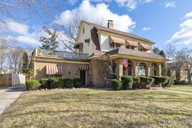 view of front of home with a gambrel roof, a balcony, a chimney, a front yard, and brick siding