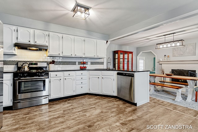 kitchen with under cabinet range hood, stainless steel appliances, a peninsula, light wood-type flooring, and dark countertops