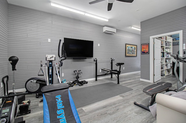 exercise room with a wall unit AC, wood finished floors, and a ceiling fan