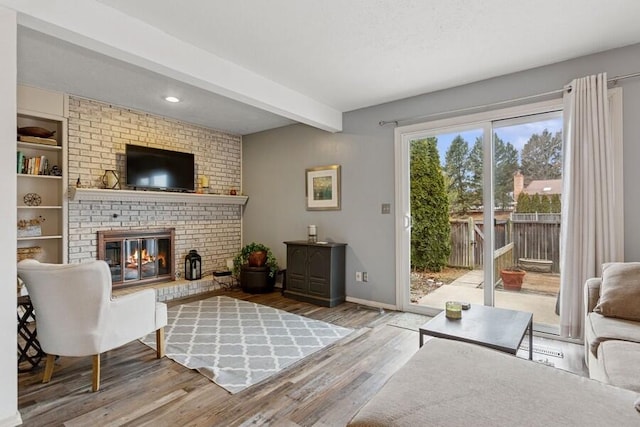 living area featuring baseboards, beamed ceiling, wood finished floors, a textured ceiling, and a brick fireplace