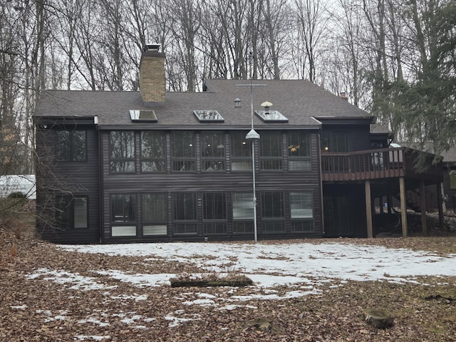 snow covered back of property with a deck, roof with shingles, a chimney, and a sunroom