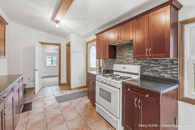 kitchen featuring light tile patterned floors, white range with gas cooktop, dark countertops, under cabinet range hood, and backsplash