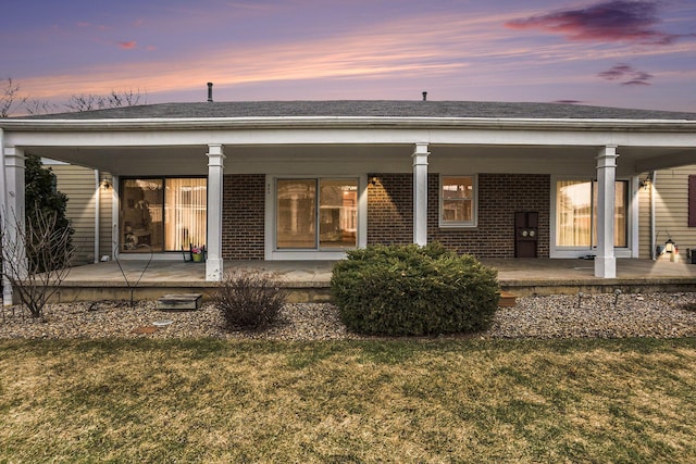 back of house at dusk featuring covered porch, a shingled roof, a yard, and brick siding