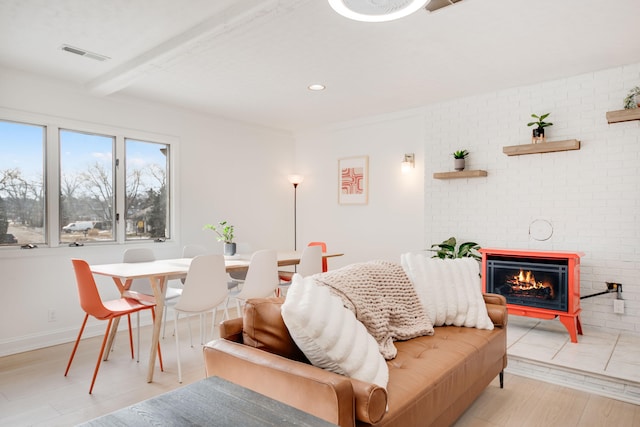 living area featuring beam ceiling, visible vents, light wood-style floors, a brick fireplace, and baseboards