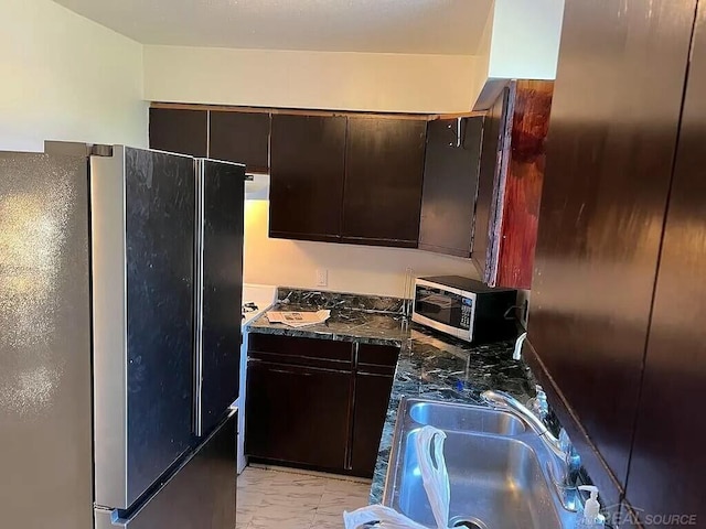 kitchen with stainless steel microwave, freestanding refrigerator, marble finish floor, dark brown cabinets, and a sink