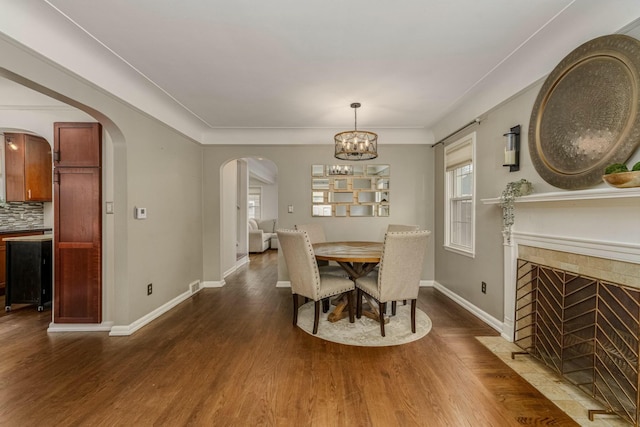 dining space featuring arched walkways, dark wood finished floors, a tiled fireplace, a chandelier, and baseboards