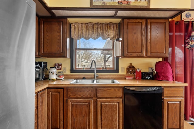 kitchen featuring black dishwasher, brown cabinetry, freestanding refrigerator, light countertops, and a sink