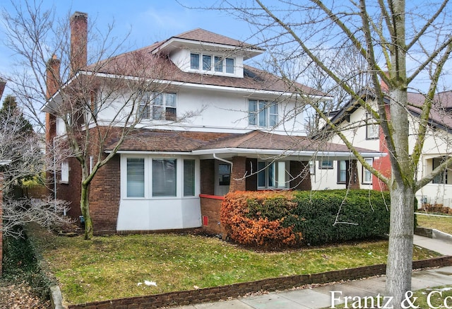 american foursquare style home featuring a front yard, brick siding, roof with shingles, and stucco siding