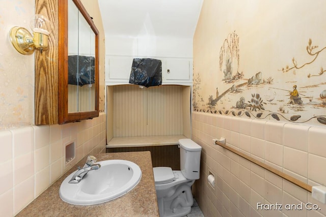 half bath featuring a wainscoted wall, wallpapered walls, a sink, tile walls, and toilet