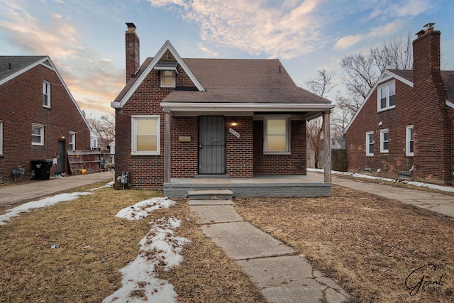 bungalow-style house featuring covered porch, brick siding, a chimney, and a shingled roof