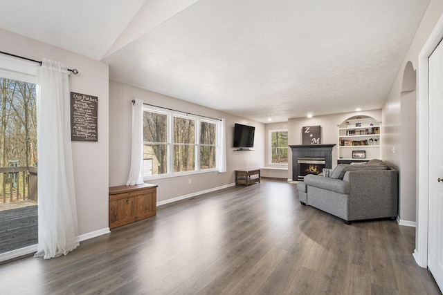 living room with baseboards, built in shelves, dark wood-style flooring, and a warm lit fireplace