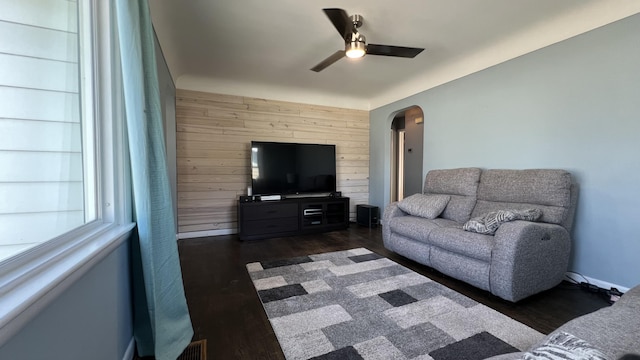 living room featuring wooden walls, a ceiling fan, dark wood-style floors, arched walkways, and an accent wall