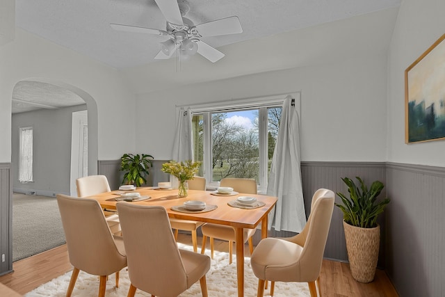 dining space featuring ceiling fan, a wainscoted wall, light wood-type flooring, arched walkways, and a textured ceiling