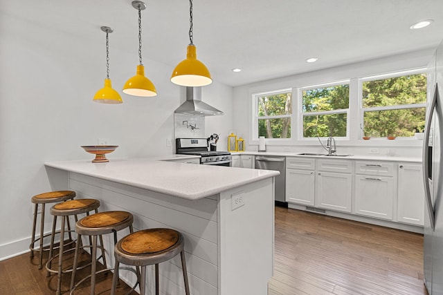 kitchen featuring a sink, stainless steel appliances, a peninsula, wall chimney range hood, and dark wood-style flooring