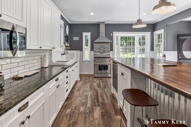 kitchen with a sink, butcher block countertops, stainless steel appliances, white cabinetry, and wall chimney range hood